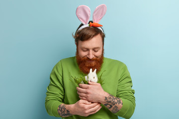 Photo of good looking man with trendy hairstyle, ginger beard, feeds little white bunny with green grass, likes domestic animals, models over blue wall. People, pet, care concept. Happy Easter