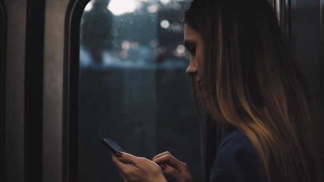Young brunette woman going somewhere in moving subway train. Girl using smartphone standing near the window on sunset.