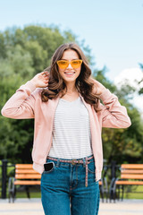 pretty girl in sunglasses smiling while touching hair in park