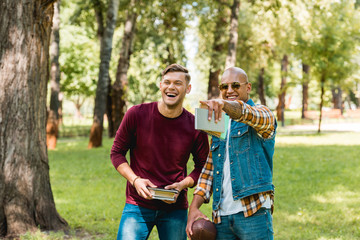 Fototapeta premium happy african american student pointing with finger and laughing while standing near handsome friend with books