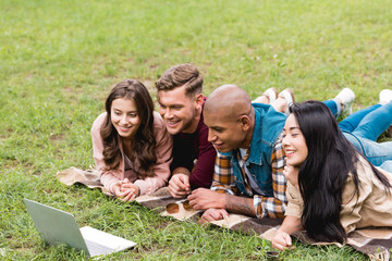 Fototapeta premium happy multicultural friends lying on blanket and looking at laptop in park