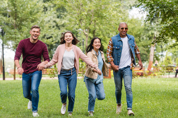 cheerful multicultural friends holding hands and running in park