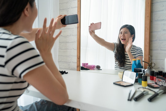 Image of happy young asian woman at home indoors sitting at dressing table doing make up with cosmetics. cheerful girl having video phone call get ready in the morning cheerful waving hand cellphone