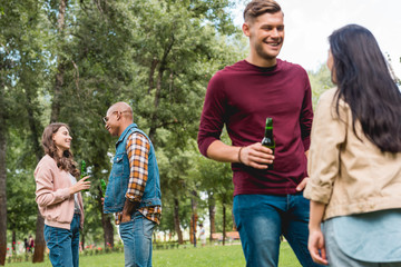 Fototapeta premium happy multicultural friends talking while holding bottles with beer in park