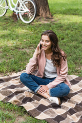 cheerful girl talking on smartphone while sitting on checkered blanket in park