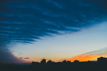 Cityscape with wonderful varicolored vivid dawn. Amazing dramatic blue cloud sky above dark silhouettes of city building roofs. Atmospheric background of orange sunrise in overcast weather. Copy space