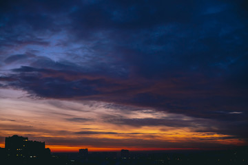 Obraz premium Cityscape with vivid warm dawn. Amazing dramatic blue violet cloudy sky above dark silhouettes of city buildings. Orange sunlight. Atmospheric background of sunrise in overcast weather. Copy space.