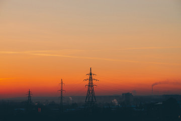 Power lines in city on dawn. Silhouettes of urban buildings among smog on sunrise. Cables of high voltage on warm orange yellow sky. Power industry at sunset. City power supply. Mist urban background.