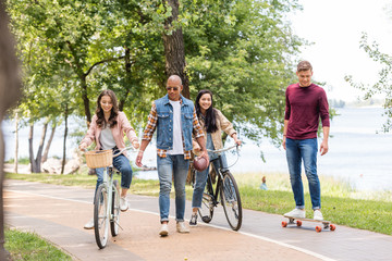 handsome african american man walking near pretty girls riding bicycles and cheerful friend longboarding in park