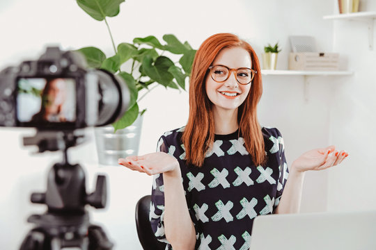 Young, redhead female fashion blogger recording video at home studio using camera with a tripod. Online tutorial, vlog, social media, personal shoper, beauty and fashion tricks.