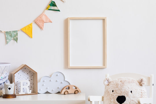 Stylish scandinavian child room with mock up photo poster frame on the white wall. Cute modern interior of nursery with boxes, teddy bear, toys.  wooden accessories and colorful flags. Real photo.