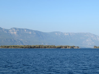 Sea coastline with mountain shore and rocky island covered with forest, view from the boat. Picturesque seascape with deep blue water in sunny day, beautiful panorama for background