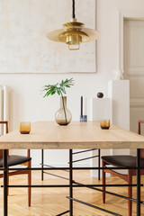 Eclectic and elegant dining room interior with design sharing table, chairs, gold pedant lamp, abstract paintings and stylish accessories. Tropical leafs in vase. Eclectic decor. Brown wooden parquet.
