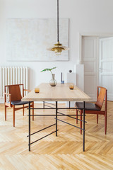 Eclectic and elegant dining room interior with design sharing table, chairs, gold pedant lamp, abstract paintings and stylish accessories. Tropical leafs in vase. Eclectic decor. Brown wooden parquet.