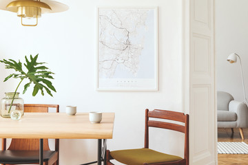 Stylish and eclectic dining room interior with mock up poster map, sharing table design chairs, gold pedant lamp and elegant sofa in second space. White walls,  wooden parquet. Tropical leafs in vase.