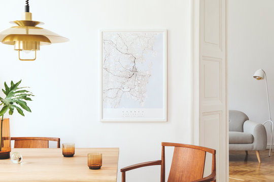 Stylish and eclectic dining room interior with mock up poster map, sharing table design chairs, gold pedant lamp and elegant sofa in second space. White walls,  wooden parquet. Tropical leafs in vase.