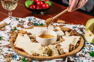 Cheese plate and chef hand wich pours honey: Sulguni, Guda, cheddar, gouda, camembert, and other served with white wine, honey and nuts on wooden board on light background. Tasty appetizers.