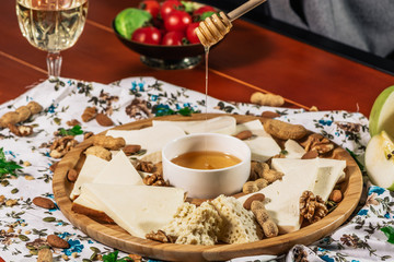 Cheese plate and chef hand wich pours honey: Sulguni, Guda, cheddar, gouda, camembert, and other served with white wine, honey and nuts on wooden board on light background. Tasty appetizers.