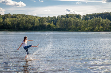 summer tourism and travel, landscape by the river. a teenage girl in a white t-shirt with long hair in blue jeans dabbles in the water. kicked the water spray in all directions