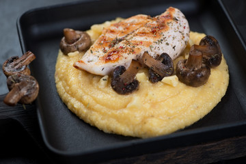 Close-up of polenta with fried chicken breast fillet and mushrooms in a cast-iron serving tray, selective focus