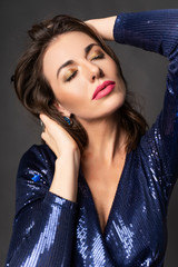 Fototapeta na wymiar A beautiful brunette woman wearing a glittery blue dress against a gray background sensually touches her hair and face with her hands. Clean, healthy skin. Advertising design.