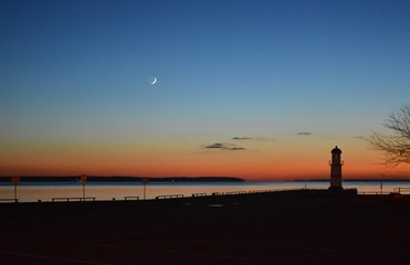 A colourful view of a sunset with a lighthouse and the moon on the shore of St-Lawrence river in Montreal, Quebec, Canada.