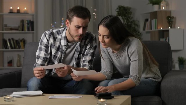 Worried couple checking bank receipts sitting on a couch in the night at home