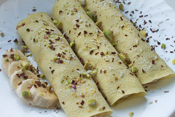 Soft focus 45 degree from above angle close up shot of three homemade crepes pancakes rolled with bananas inside topped with chocolate and cocos shavings and pistachios on a white plate.