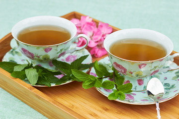 Herbal mint tea served in two china porcelain cups with bunch of fresh leaves nearby and a silver spoon in wooden tray on linen textile, copy space, close-up, alternative medicine concept