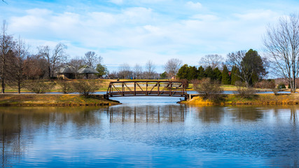 A small bridge in a public park on a calm Winter day. Light clouds are in the sky. Brown and Green in abundance