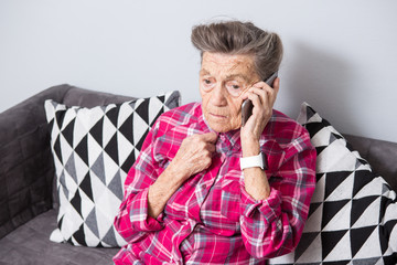 An old elderly woman grandmother with gray hair sits at home on the couch using the hand phone, a telephone conversation to hear the bad news. Emotion fear scare
