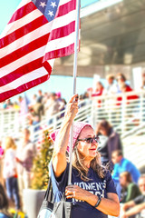 Older woman with flag and Womens March tee shirt at political rally with crowd blurred behind her