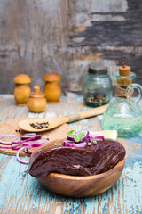 A piece of raw beef liver in a bowl, onion, lemon and spices for cooking on a wooden table