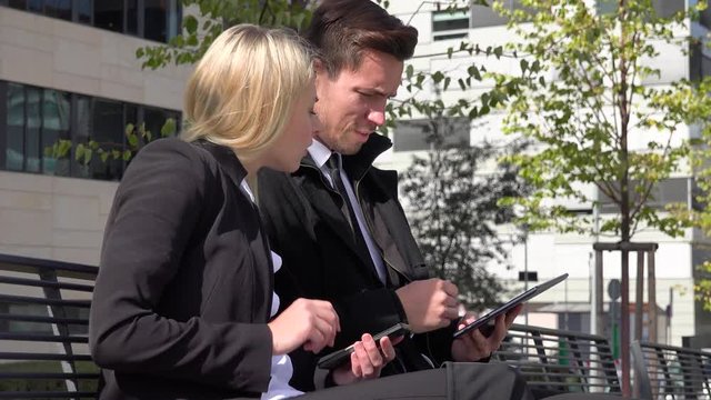 A businessman and a businesswoman sit on a bench in an urban area, work on a smartphone and a tablet and talk - office buildings in the background