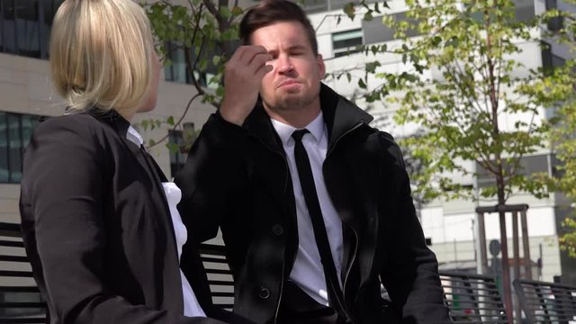 A businessman and a businesswoman sit on a bench and argue in an urban area - office buildings in the background
