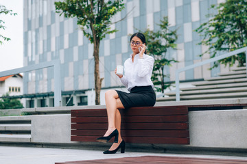 Fototapeta premium Busy Asian businesswoman in eyeglasses talking about business discussion with smartphone while drinking coffee sitting in front office building background- woman office worker- businesswoman concept