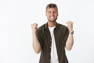Guy happy finally catching dream. Relieved and delighted attractive blond man with bristle close eyes and smiling satisfied, clench fists in triumph and victory making yes gesture over gray wall