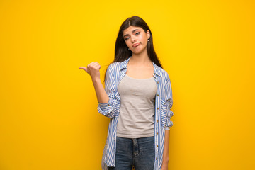 Teenager girl over yellow wall unhappy and pointing to the side