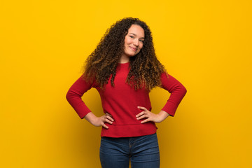 Teenager girl with red sweater over yellow wall posing with arms at hip and smiling