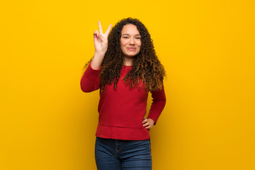 Teenager girl with red sweater over yellow wall smiling and showing victory sign