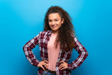 Teenager girl over blue wall posing with arms at hip and laughing looking to the front