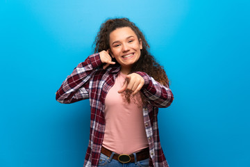 Teenager girl over blue wall making phone gesture and pointing front