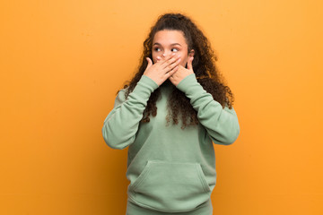 Teenager girl over ocher wall covering mouth and looking to the side