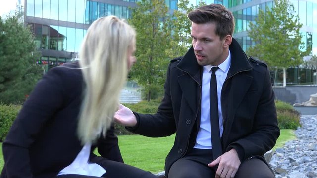 A businessman and a businesswoman sit on a bench and argue in an urban area - an office building in the background