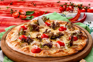 Caesar pizza with cheese, cherry tomatoes, chicken and lettuce on a round wooden board on a red wooden background, decorated with napkins, chili pepper and cherry tomatoes. close up. top view