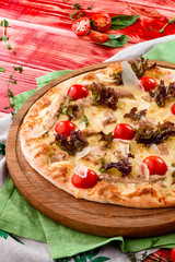 Caesar pizza with cheese, cherry tomatoes, chicken and lettuce on a round wooden board on a red wooden background, decorated with napkins, chili pepper and cherry tomatoes. close up. top view