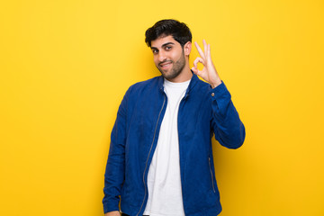 Man with blue jacket over yellow wall showing ok sign with fingers