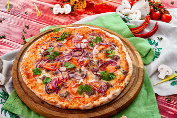 Italian Pizza with meat, tomatoes, garlic, onions and cheese on a round wooden board on a red wooden background, decorated with napkins, chili pepper and cherry tomatoes. close up. top view