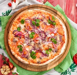 Italian Pizza with meat, tomatoes, garlic, onions and cheese on a round wooden board on a red wooden background, decorated with napkins, chili pepper and cherry tomatoes. close up. top view