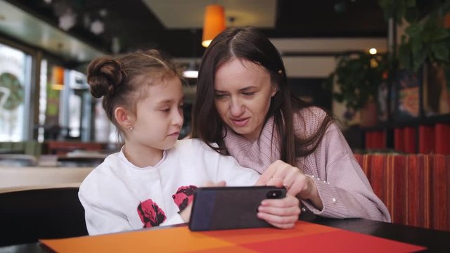 Attractive caucasian mother and her daughter using smartphone at the cafe. An advanced little girl shows and explains to his mother something on a smartphone.
